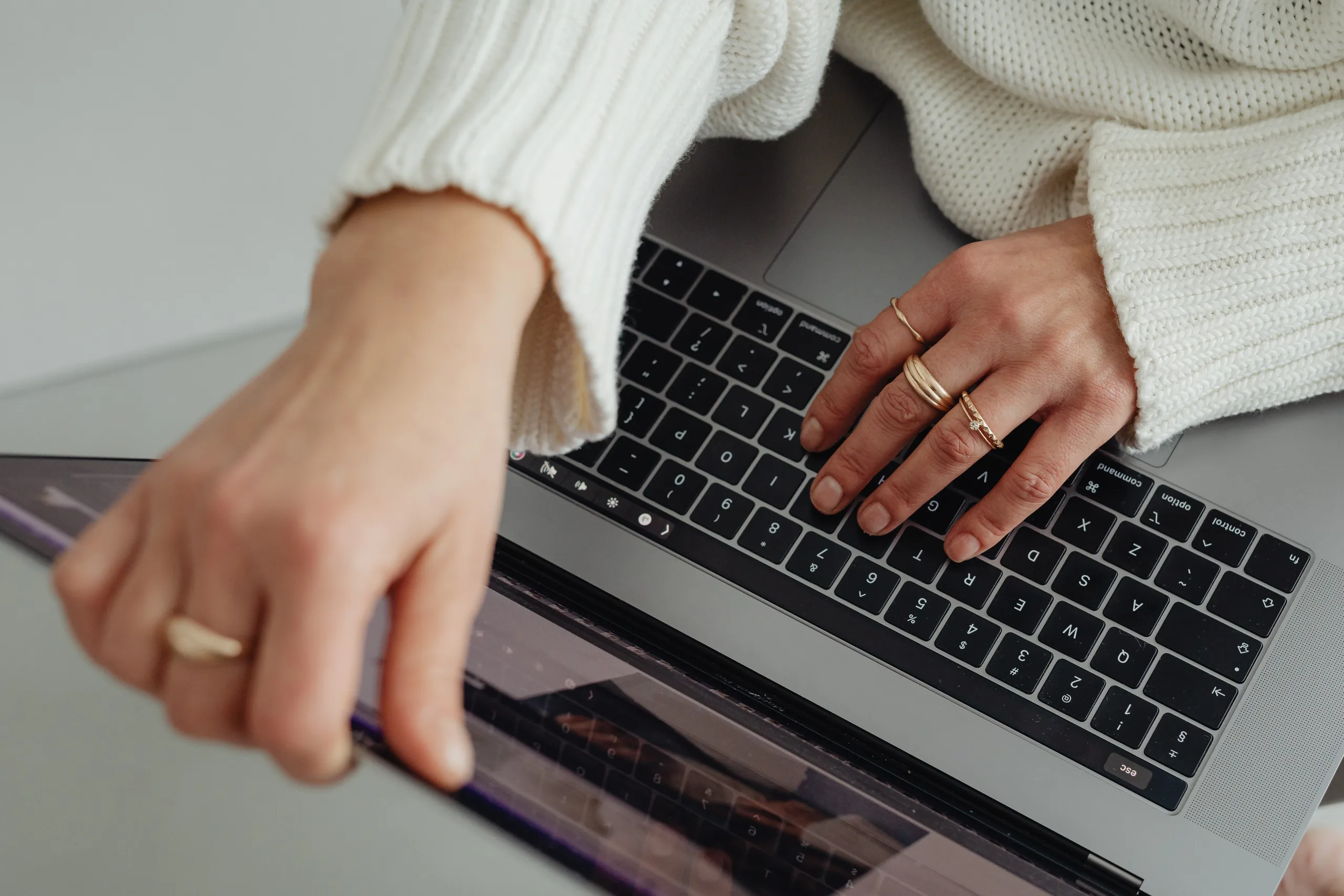 Hands typing on a laptop beside a tablet on a light gray desk.