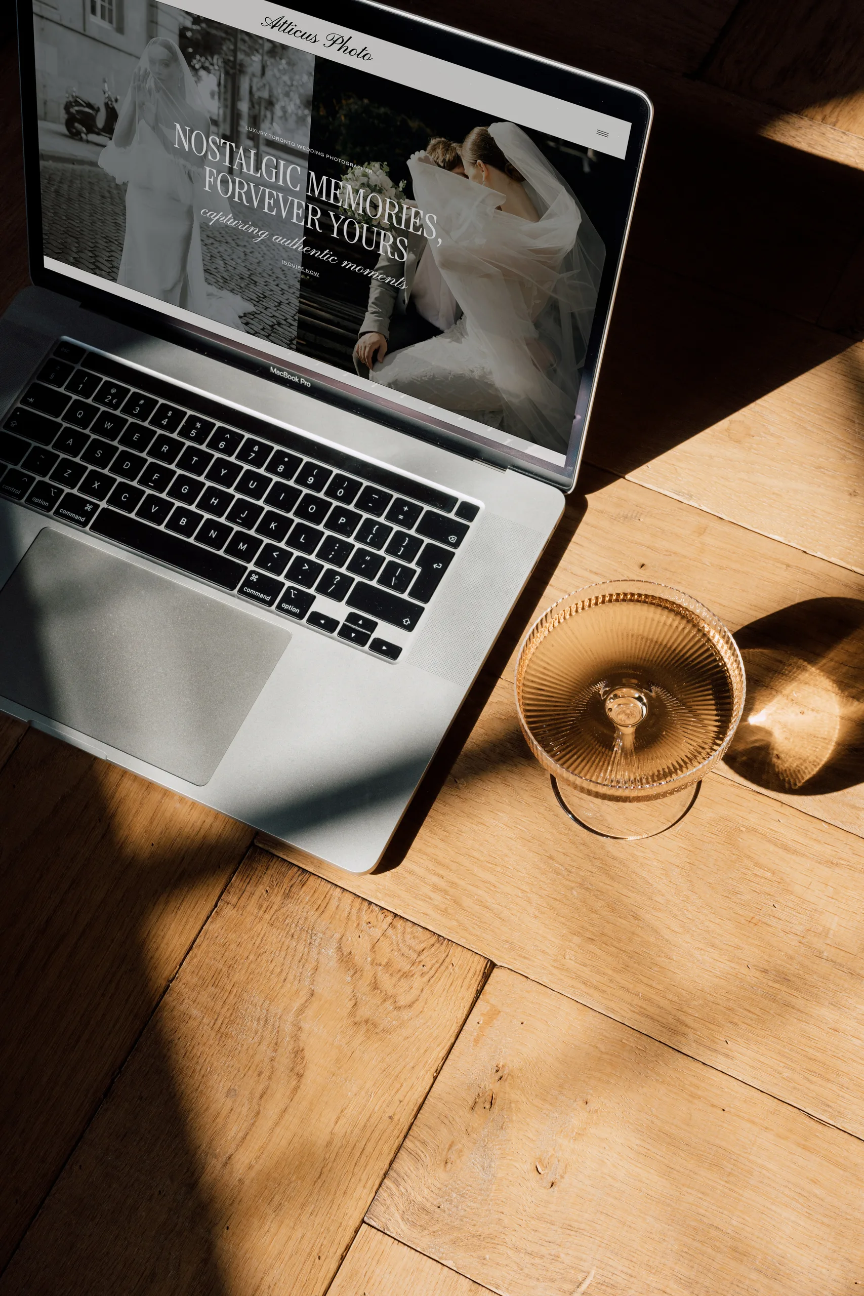Laptop on a wooden floor displays a wedding website beside a gold-rimmed glass.