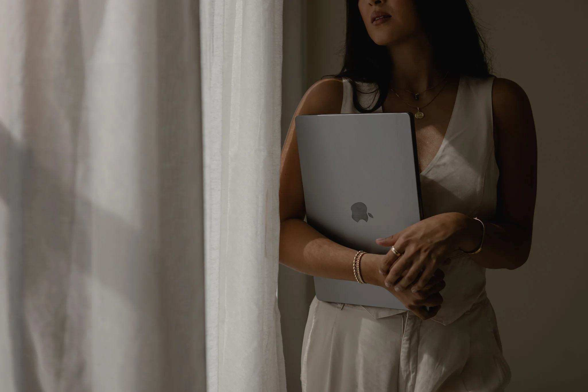 Woman in a light outfit holding a silver laptop beside white curtains.